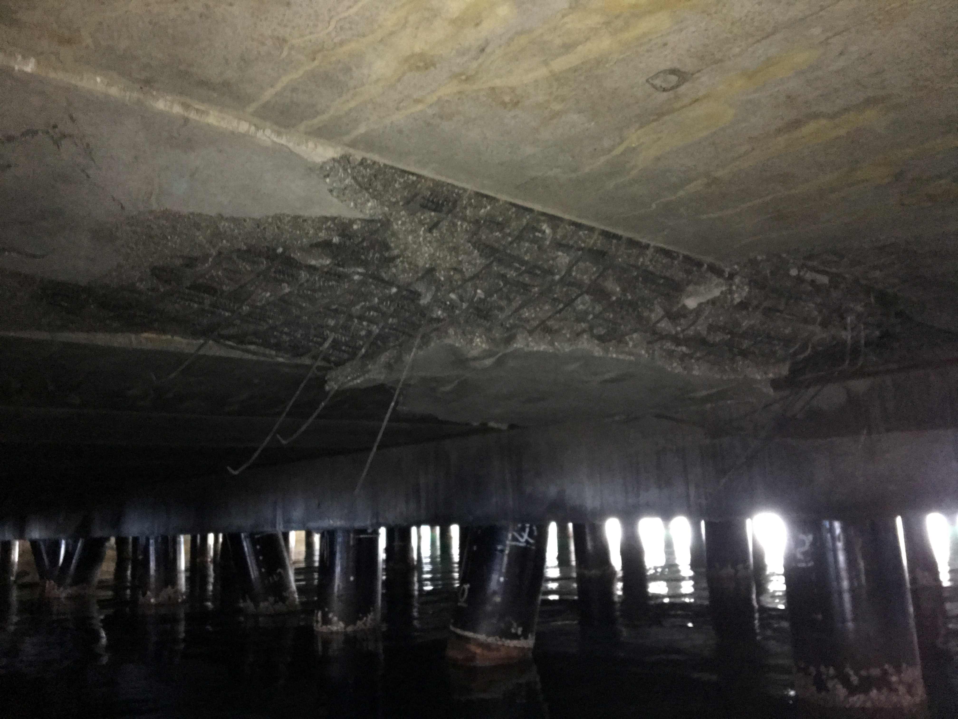 Underside of a marine wharf showing concrete deterioration and piles in water during inspection